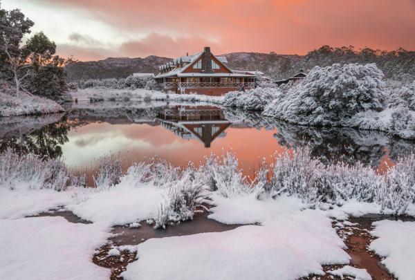 A wooden lodge surrounded by snow-covered trees reflects across a glassy lake beneath a pink-tinged sunset, Peppers Cradle Mountain Lodge, Cradle Mountain-Lake St Clair National Park, Tasmania. © Tourism Australia
