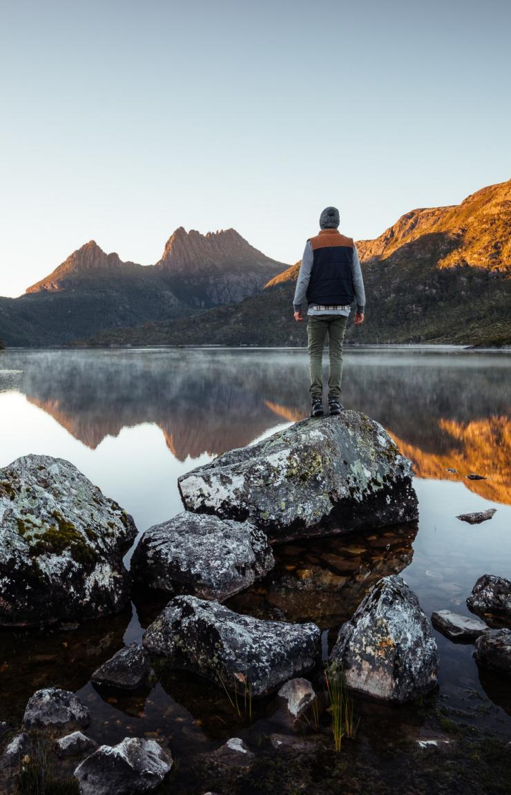 A person stands on a rock on the edge of a still lake reflecting rugged peaks illuminated by golden morning light, Cradle Mountain-Lake St Clair National Park, Tasmania. © Tourism Australia