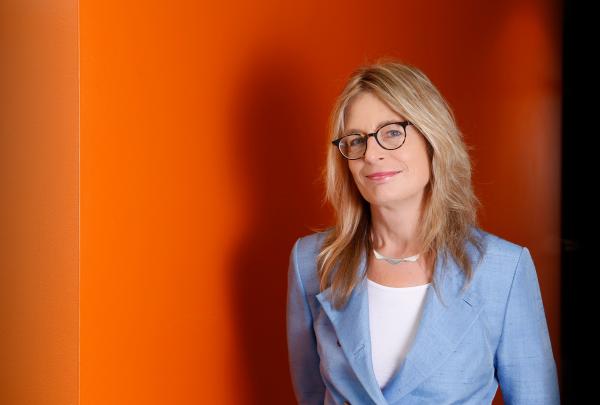 Professor Helen Christensen, Director and Chief Scientist at the Black Dog Institute, wears glasses and a light blue blazer against a vibrant orange wall. © Australia Unlimited