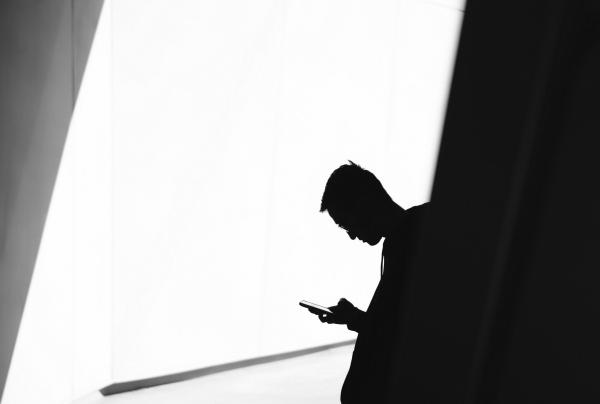 A black-and-white silhouette of a person looking down at their smartphone stands against a brightly lit geometric wall. © Warren Wong, Australia Unlimited