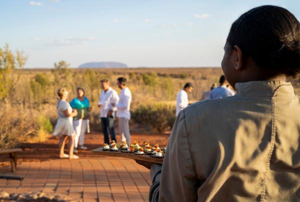 Server carries a tray of canapés towards guests socialising outdoors in golden light with Uluru in the distance during a Tali Wiru experience by Voyages Indigenous Tourism Australia, Northern Territory. © Voyages Indigenous Tourism Australia