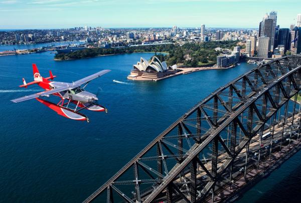 A red and white Sydney Seaplanes aircraft soars past the Sydney Harbour Bridge over clear blue waters, taking in panoramic views of the Opera House and city skyline in Sydney Harbour, New South Wales. © Sydney Seaplanes