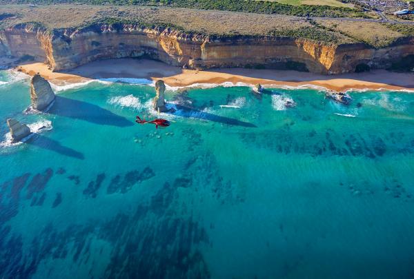 A red Microflite helicopter soars over turquoise ocean waters and limestone stack rock formations along the coastal cliffs of the Twelve Apostles on the Great Ocean Road in Victoria. © Microflite