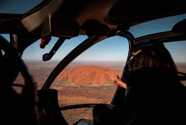 A passenger inside a helicopter points out the window at the vibrant red expanse of Uluru rising from the desert landscape during a scenic flight over Uluru-Kata Tjuta National Park, Northern Territory. © Tourism Australia