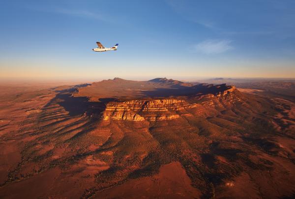 A small light aircraft takes a scenic flight over the dramatic red rock amphitheatre of Wilpena Pound in Ikara-Flinders Ranges National Park, South Australia, illuminated by warm afternoon light. © South Australian Tourism Commission