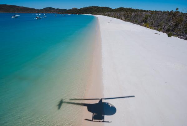 A helicopter casts its dark shadow over pure white silica sand and turquoise ocean waters along the shore of Whitehaven Beach in the Whitsundays, Queensland, with boats anchored in the distance. © Tourism and Events Queensland