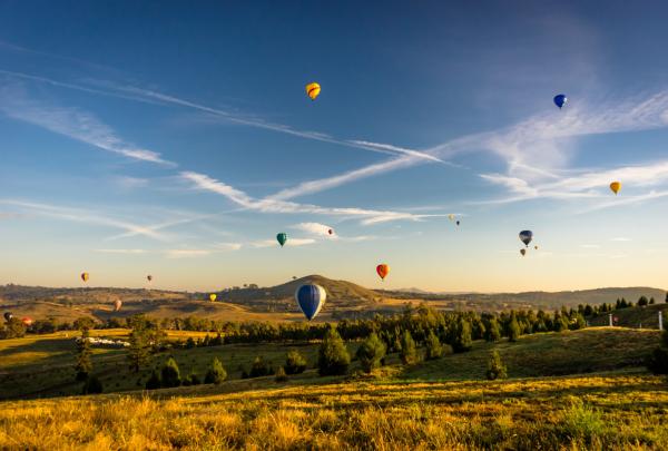 Colorful hot air balloons float across a bright blue sky streaked with vapor trails above rolling golden hills and green trees in Canberra, Australian Capital Territory at dawn. © VisitCanberra