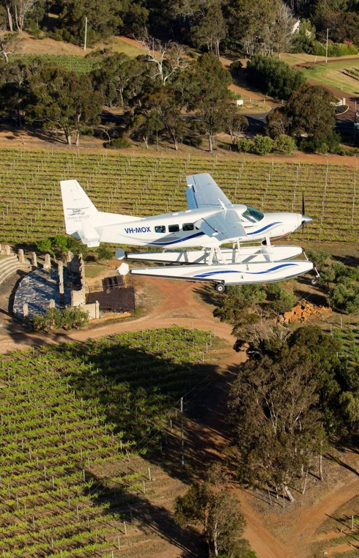 A white Swan River Seaplanes aircraft glides over lush green vineyard rows and rolling countryside in the Margaret River region, Western Australia, under bright daytime sunlight. © Swan River Seaplanes