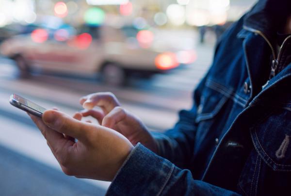 A person in a denim jacket types on a Smartphone at dusk, with blurred city traffic and soft streetlights in the background. © Getty Images