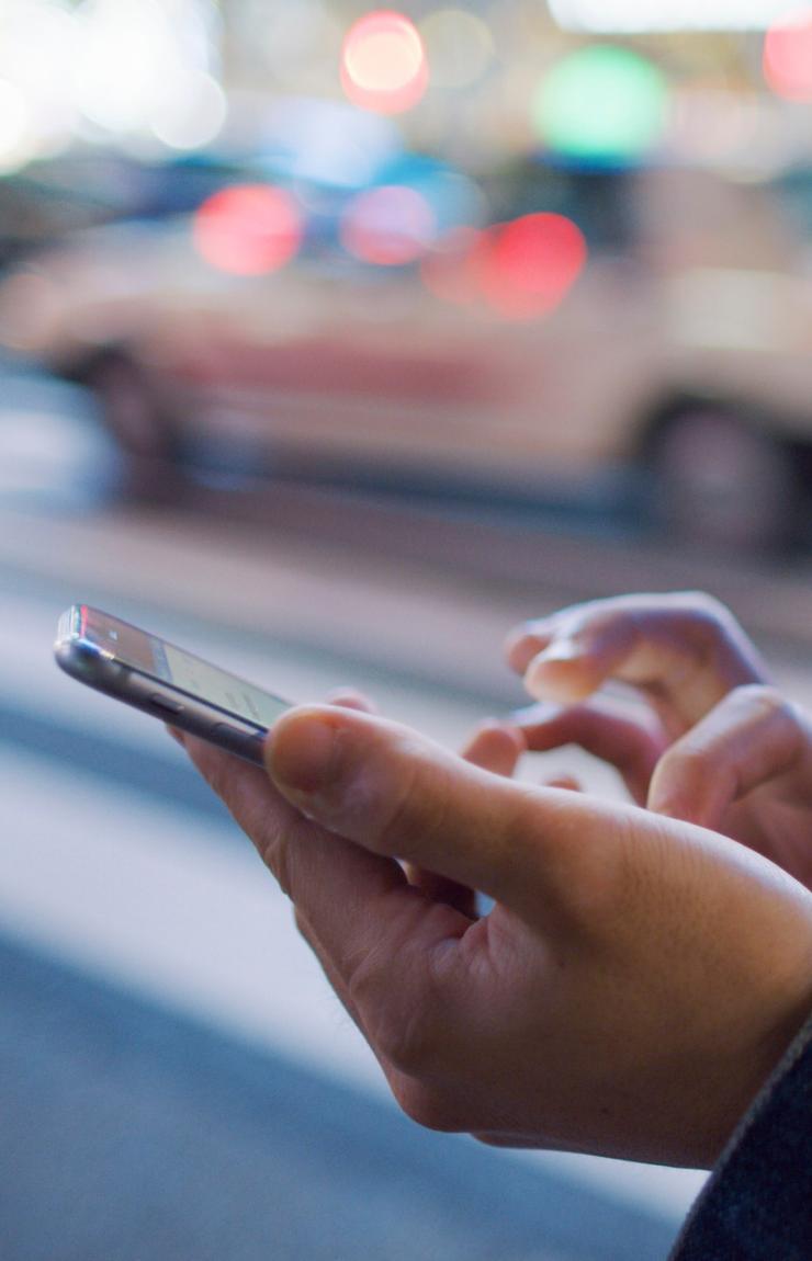 A person in a denim jacket types on a Smartphone at dusk, with blurred city traffic and soft streetlights in the background. © Getty Images