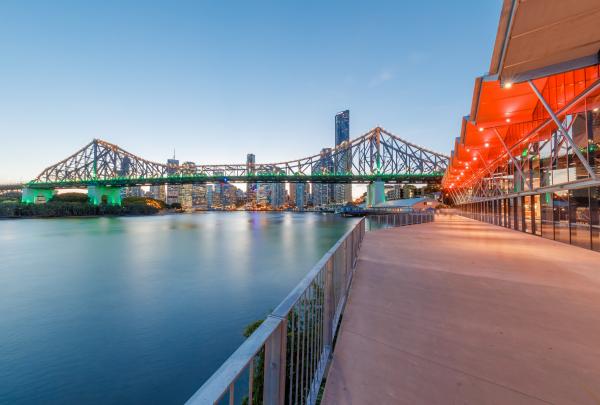 Howard Smith Wharves boardwalk in Brisbane, Queensland glowing at dusk alongside the calm river, with orange canopy lights shining and the illuminated Story Bridge and city sky scrapers beyond. © Howard Smith Wharves