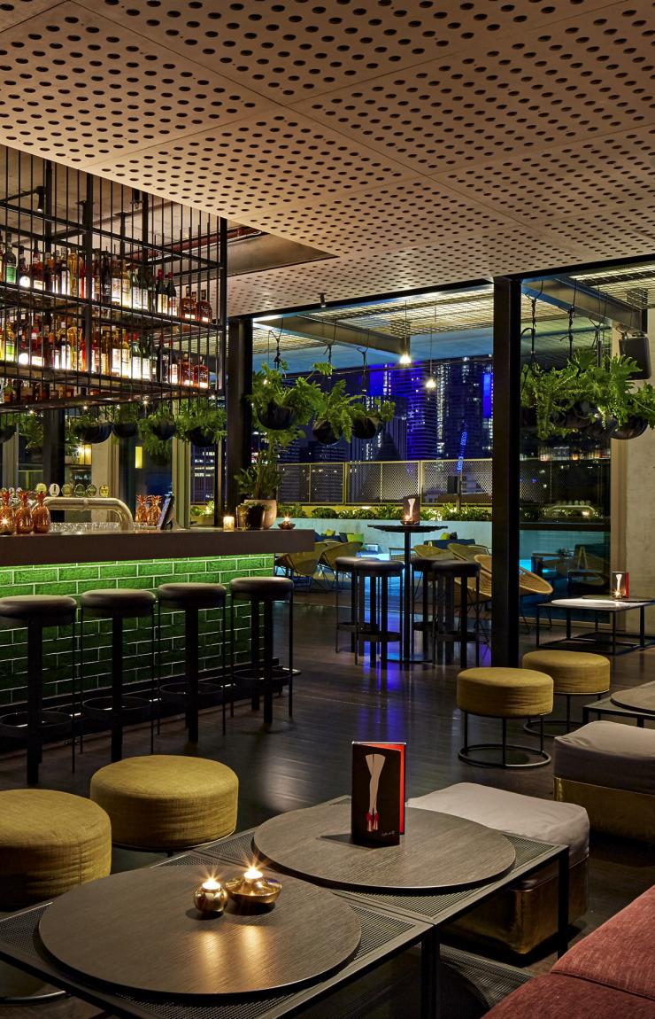 A green tiled bar illuminated at night with warm mood lighting and low seating at Rooftop at QT, Melbourne, Victoria, offering views of the city skyline through large open-air glass windows. © QT Melbourne