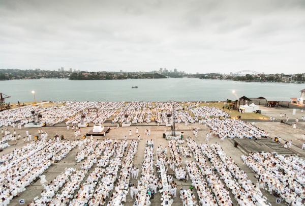 An expansive high-angle view captures thousands of guests dressed in white dining at long outdoor tables along the waterfront, Cockatoo Island, Sydney Harbour, New South Wales. © Sydney Harbour Federation Trust