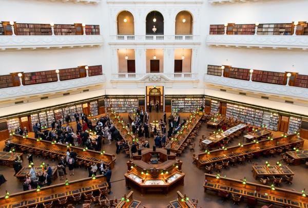 An expansive high-angled view reveals octagonal wooden reading desks with green lamps set beneath multi-tiered balconies lined with books, La Trobe Reading Room, State Library of Victoria, Melbourne, Victoria. © Showtime Event Group and @cassandraladru