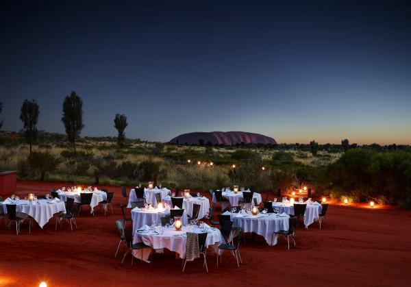 Candlelit dining tables set on red desert soil glow under a twilight sky with views of Uluru in the distance, Ayers Rock Resort, Uluru-Kata Tjuta National Park, Northern Territory. © Ayers Rock Resort