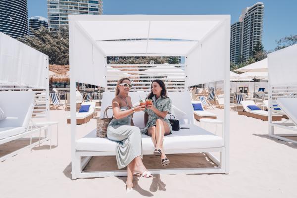 Two women toast cocktails while relaxing on a luxurious white beach cabana on the sand against a backdrop of coastal high-rises, Kurrawa Beach Club, Brisbane, Queensland. © Kurrawa Beach Club