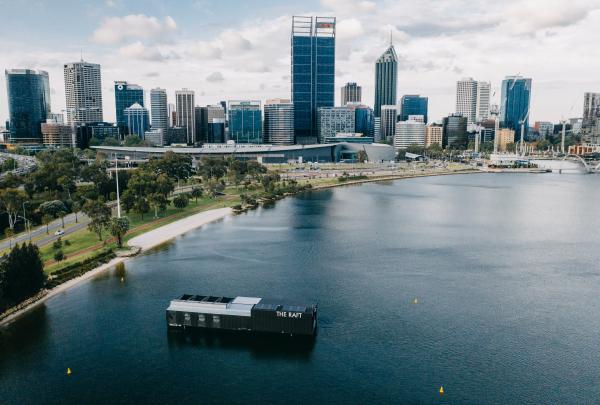An aerial view showcases a floating venue anchored on the river with city skyscrapers in the background, The Raft Perth, Western Australia. © Shot by Thom