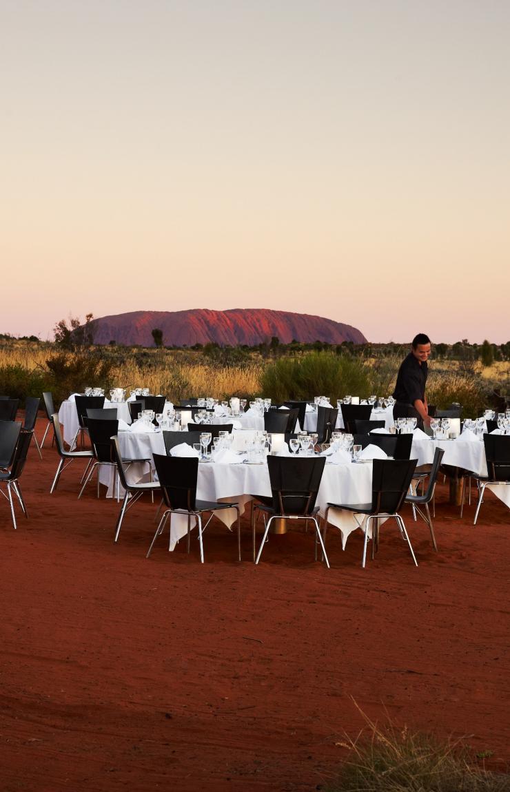 Elegant white-cloth dining tables set on red desert sand offer an open-air dinner experience with views of Uluru at sunset, Tali Wiru, Uluru-Kata Tjuta National Park, Northern Territory. © Tourism Australia