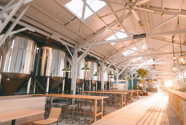 Sunlight streams into the spacious industrial venue at Gage Roads Brew Co., Fremantle, Western Australia, highlighting high white rafters, large shiny brewing tanks, and long wooden dining tables. © Gage Roads Brew Co.