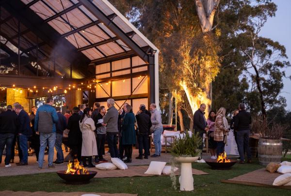 Guests socialise outdoors at dusk at Lot 100 in the Adelaide Hills, South Australia, gathering around warm fire pits and string lights outside a modern industrial barn amidst towering gum trees. © Lot 100