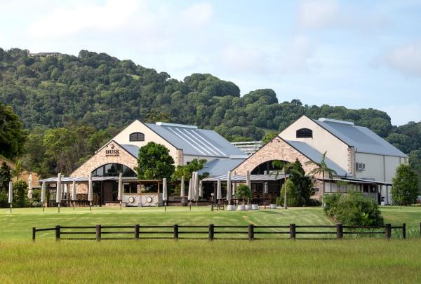 The picturesque countryside grounds and modern farm-style distillery buildings of Husk in North Tumbulgum, New South Wales, set against rolling green hills beneath a soft light blue sky. © Husk
