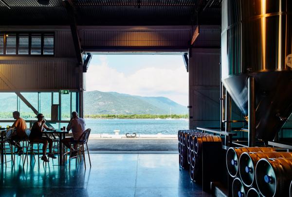 Patrons relaxing inside Hemingway’s Brewery in Port Douglas, Queensland, looking past large shiny brewing tanks and steel kegs out through open barn doors to vibrant blue water and coastal hills. © Hemingway’s Brewery