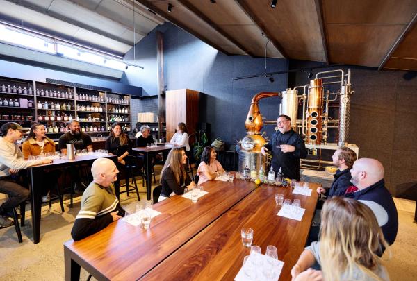 Guests gather around long timber tables for a masterclass hosted next to shining copper stills inside Four Pillars Gin, Yarra Valley, Victoria, taking in the warm atmosphere. © Tourism Australia 