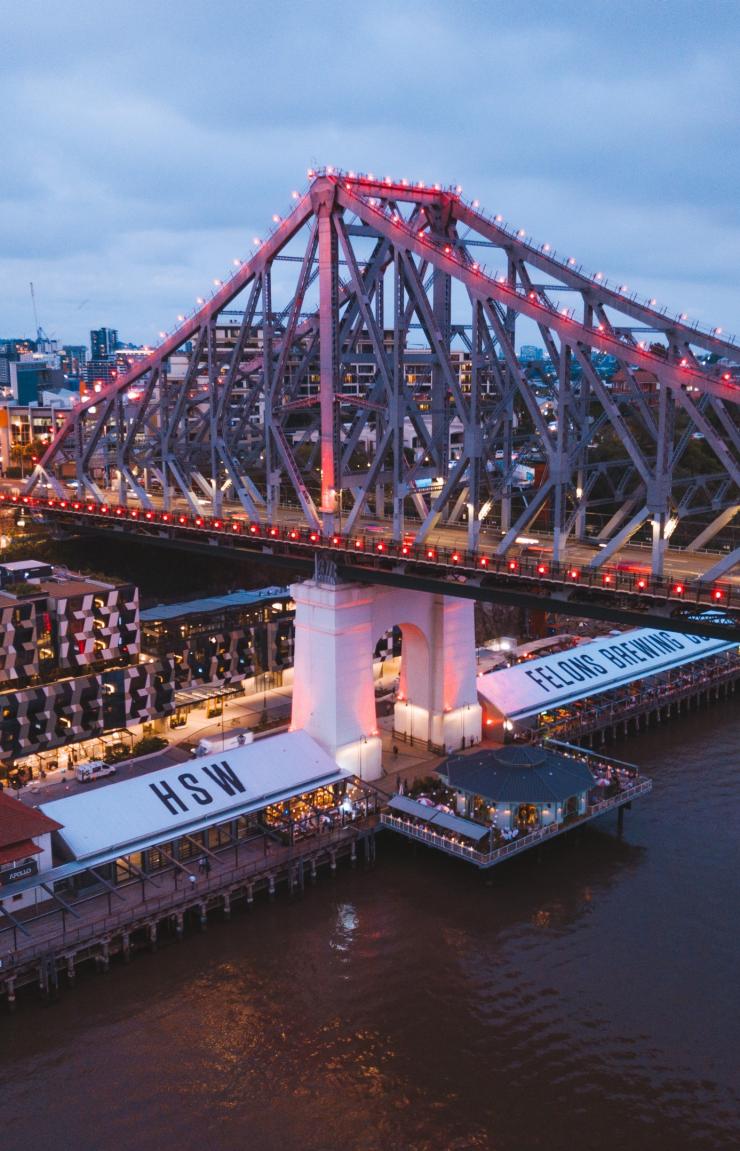 Dusk falls over Howard Smith Wharves in Brisbane, Queensland, where the illuminated Story Bridge casts a warm reddish glow above riverside venues along the calm waters of the Brisbane River. © Tourism and Events Queensland