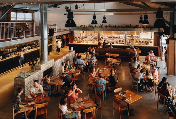 Diners enjoy meals at warm wooden tables in the spacious industrial-style dining room of Three Blue Ducks at URBNSURF, Melbourne, Victoria, lit by low pendant lights near an open kitchen. © URBNSURF