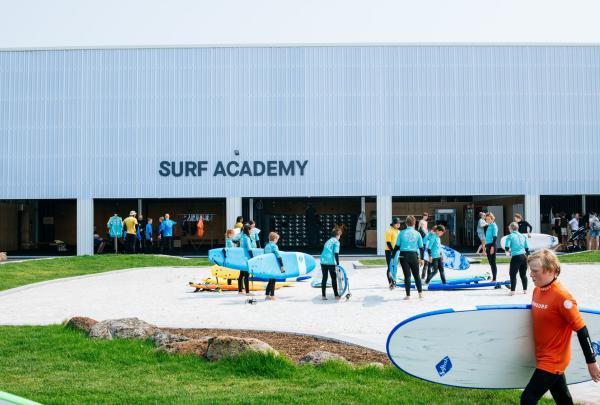 Surfers carry surfboards across a sandy patch outside the modern Surf Academy building during a group lesson at URBNSURF, Melbourne, Victoria, with participants gathering on grassy lawns nearby. © URBNSURF