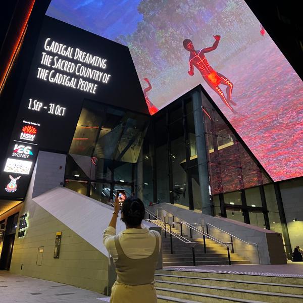 A person takes a photo on their phone of a large digital screen displaying First Nations artwork above the entrance outside ICC Sydney, Sydney, New South Wales. © Tourism Australia