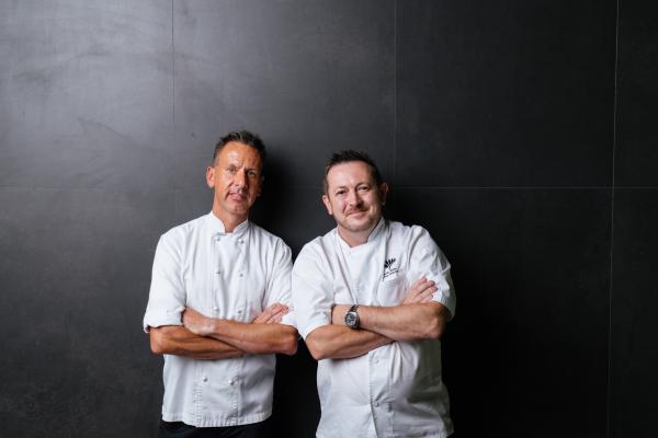 Two smiling chefs in white uniforms stand side-by-side with folded arms against a dark backdrop at the Perth Convention and Exhibition Centre, Perth, Western Australia. © Fresh Culinary Direction