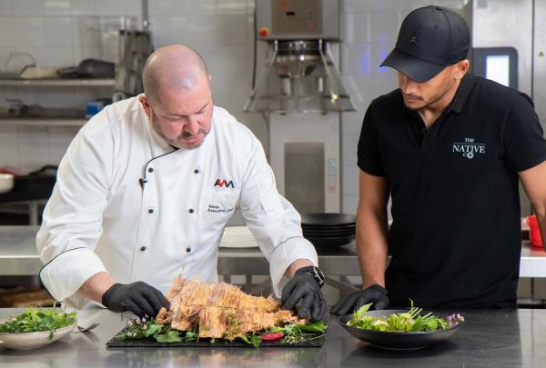 A chef in a white uniform carefully plates a dish with native greens while a colleague in a black cap watches in a commercial kitchen at Adelaide Convention Centre, Adelaide, South Australia. © Tourism Australia