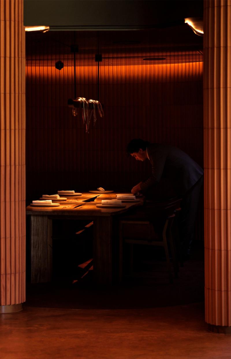At Yiaga, Melbourne, Victoria, warm light illuminates a moody dining space lined with fluted timber panels. In the background, a staff member meticulously sets a long wooden table beneath soft, atmospheric lighting. © Yiaga
