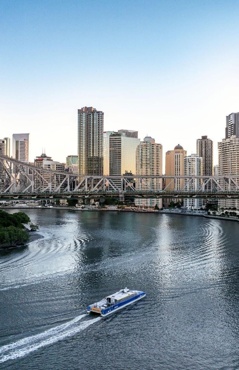 A blue and white CityCat ferry cruises along the wide Brisbane River in Queensland, passing under the historic steel-truss Story Bridge. High-rise city skyscrapers line the riverbank under a clear, pale blue sky. © Tourism Australia