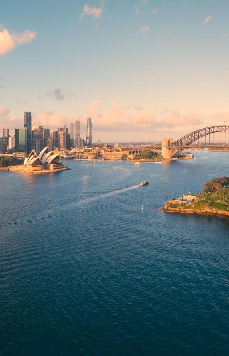 A sweeping panoramic aerial view captures the idyllic and iconic Sydney Harbour in the soft glow of early morning sunshine. The image stretches from the skyscrapers of the CBD and the lush Botanic Gardens across to the famous Sydney Opera House and Harbour Bridge. North Sydney and Kirribilli are clearly visible, basking in the golden light, with the calm harbour waters reflecting the warm hues of the sunrise. © Tourism Australia