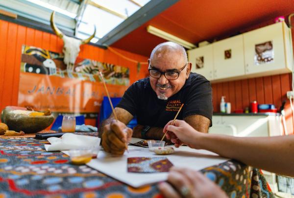 At Janbal Gallery, Mossman Gorge, Tropical North Queensland, a smiling Indigenous artist wearing glasses guides a visitor through a traditional dot painting workshop at a colourful patterned table using brushes and small canvases. © Tourism Australia