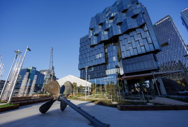 At 1 Hotel Melbourne, Melbourne, Victoria, a large, weathered steel ship propeller sculpture sits in an open paved plaza area. Behind it, the striking geometric blue glass facade of the contemporary hotel towers into the clear blue sky. ©Tourism Australia