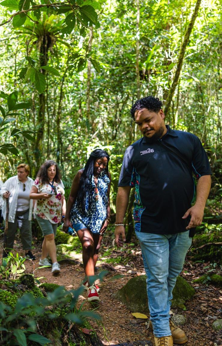 At Mossman Gorge Cultural Centre, Mossman Gorge, Tropical North Queensland, an Indigenous tour guide leads a diverse group of visitors along a dirt walking track that winds through a dense, lush and sun-dappled ancient rainforest. © Tourism Australia