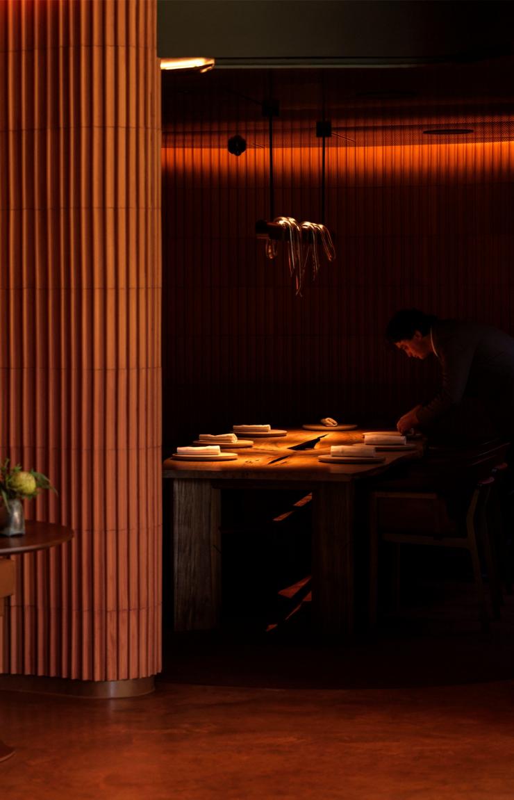 At Yiaga, Melbourne, Victoria, warm light illuminates a moody dining space lined with fluted timber panels. In the background, a staff member meticulously sets a long wooden table beneath soft, atmospheric lighting. © Yiaga