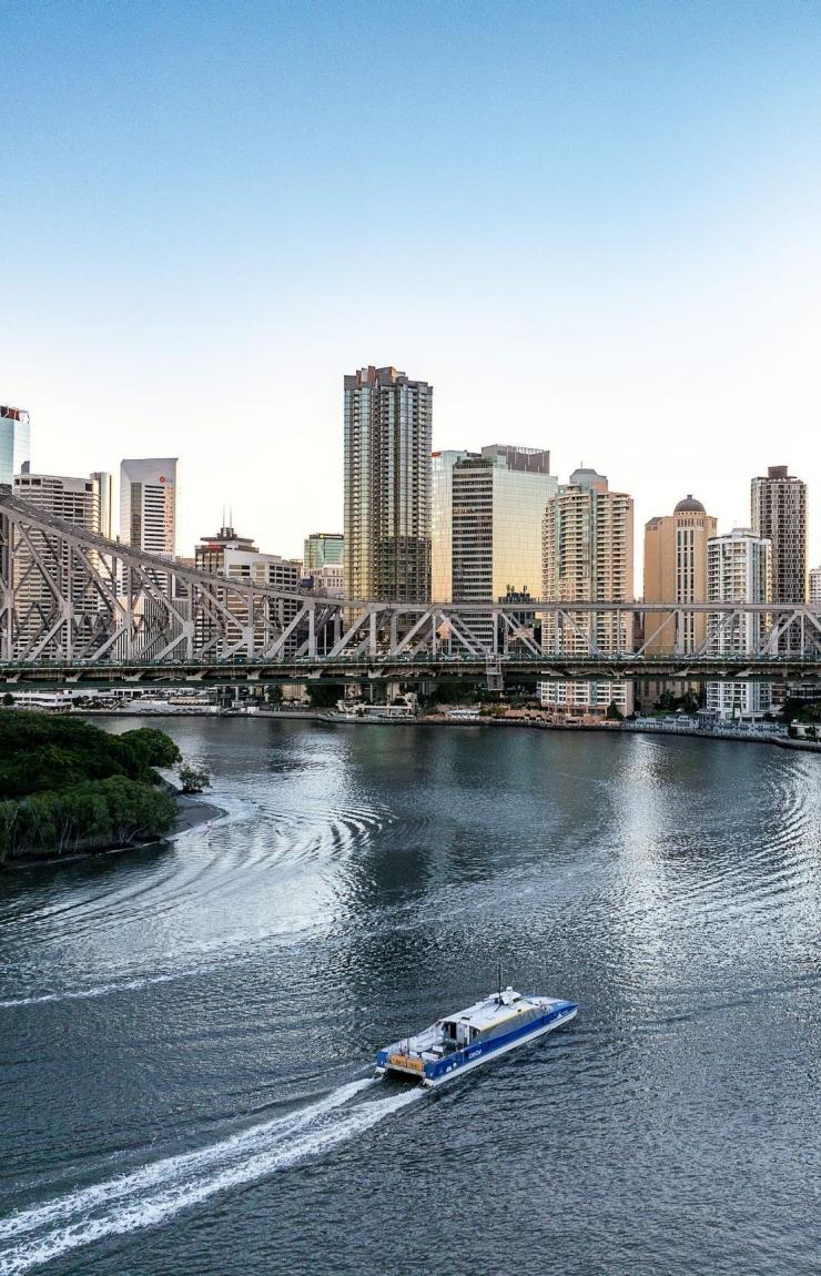 A blue and white CityCat ferry cruises along the wide Brisbane River in Queensland, passing under the historic steel-truss Story Bridge. High-rise city skyscrapers line the riverbank under a clear, pale blue sky. © Tourism Australia