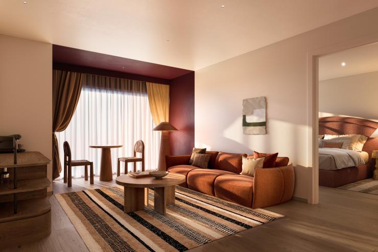The stylish hotel lounge room at Hyde Perth in Western Australia features a plush terracotta sofa, a striped area rug, a round timber coffee table, and a small dining setting by the window. An open doorway reveals the main bedroom. © Hyde Perth