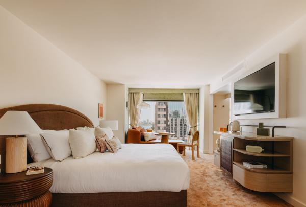 A modern, warm-toned room at Hyde Perth in Western Australia features a king bed with white linen, a curved brown headboard, a large wall-mounted TV, and a window seating area with a city view framed by neutral curtains. © Ridwaan Moolla & Melissa Mills