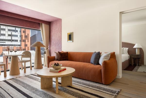 A stylish guest suite living area at Hyde Perth, Western Australia, features an earthy orange lounge, wooden coffee table, striped rug, a pink accent wall framing city views, and an open doorway leading to a cosy bedroom. © Jenna French
