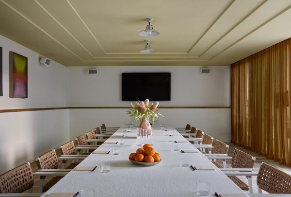 A stylish boardroom at Hyde Perth, Western Australia, features a long table with a white tablecloth, woven-style armchairs, a bowl of fresh oranges, a vase of native flowers, a sleek wall-mounted TV, and a stepped mustard ceiling. © Jenna French