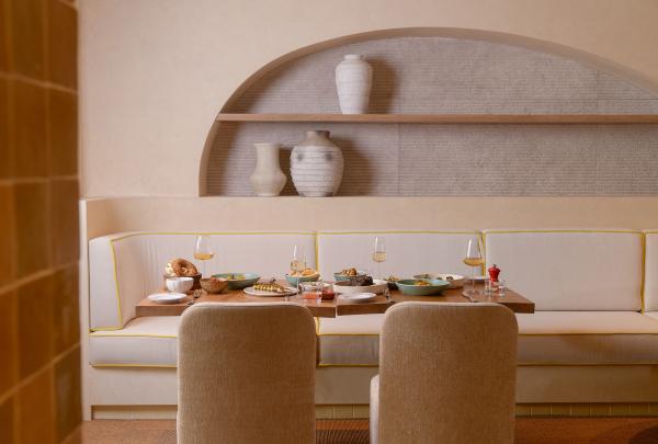 An elegant dining setting at Hyde Perth in Western Australia features a timber table spread with dishes and wine glasses, set against a cream banquette with yellow piping and a curved wall recess displaying ceramic vases. © Ryan Cubbage