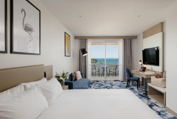 A bright and modern guest room at voco Darwin Suites in Darwin, Northern Territory, featuring a plush white bed, blue lounge, desk and patterned carpet. Sliding glass doors open to a private balcony with beautiful ocean views. © voco Darwin Suites