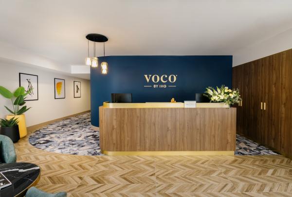 The welcoming reception area at voco Darwin Suites in Darwin, Northern Territory, features a curved timber desk with gold trim set against a deep blue feature wall displaying the hotel logo, alongside herringbone timber flooring. © voco Darwin Suites