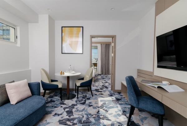 The stylish, modern living area at voco Darwin Suites in Darwin, Northern Territory, features a curved blue lounge, a small round dining table with two chairs, and a study desk, set on a vibrant blue and grey patterned carpet. © voco Darwin Suites