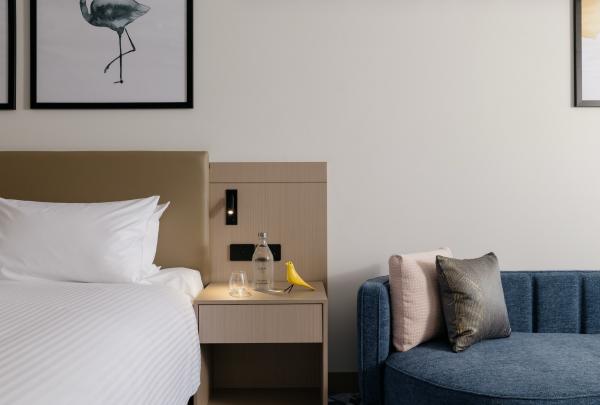 A contemporary room detail at voco Darwin Suites in Darwin, Northern Territory, showcases a crisp white bed, a light timber bedside table topped with a playful yellow bird figurine, and a curved blue lounge with elegant cushions. © voco Darwin Suites